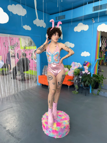 Tattooed model with short curly hair wearing a shimmery pink bodysuit featuring a blue butterfly in the center. Model is wearing fuzzy pink antenna 