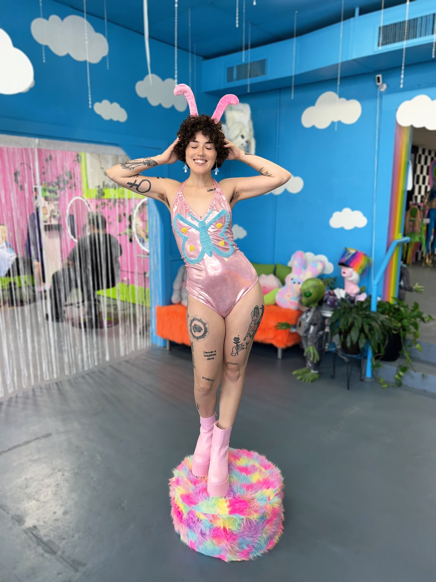 Tattooed model with short curly hair wearing a shimmery pink bodysuit featuring a blue butterfly in the center. Model is wearing fuzzy pink antenna 
